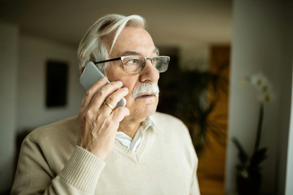 An elderly man with white hair and glasses, wearing a light sweater, holds a smartphone to his ear, appearing thoughtful as he looks into the distance indoors—perhaps considering life care planning in the Twin Cities.