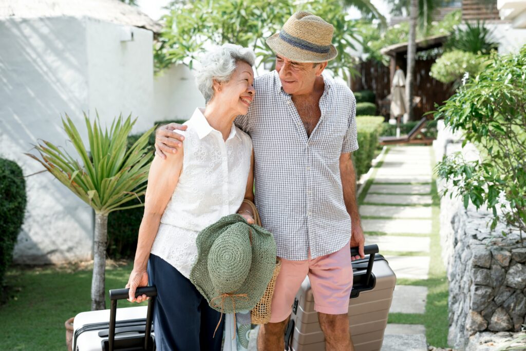 An elderly couple smiles at each other while walking outdoors with suitcases. Dressed for summer and holding a sun hat, they look ready for vacation—capturing the joy and independence Minnesota elder law helps seniors preserve.