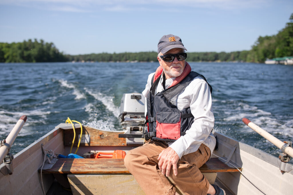 An older man wearing sunglasses, a cap, and a life jacket steers a small motorboat on a lake near the Twin Cities, with trees and blue sky in the background and water trailing behind the boat.