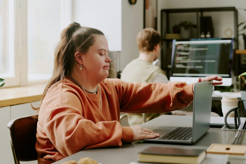 A young woman with Down syndrome is using a laptop at a desk in a bright Twin Cities office. She reaches out to open or adjust the screen while another person works at a computer in the background.