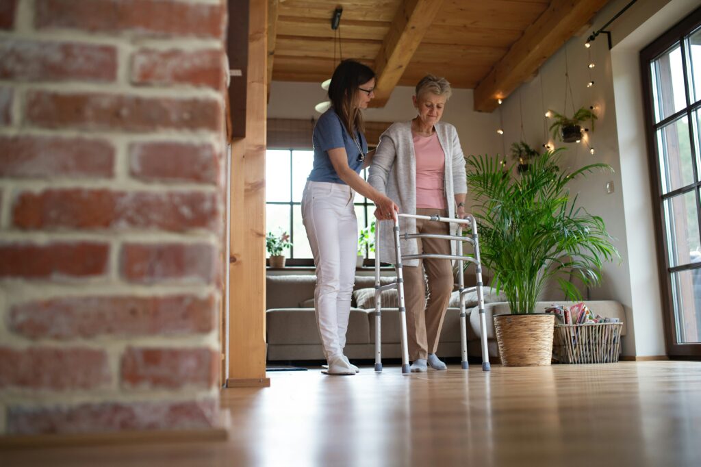 A young woman assists an older woman using a walker in a cozy, sunlit living room with wooden ceilings, a brick wall, plants, and string lights—offering support often seen in guardianships.