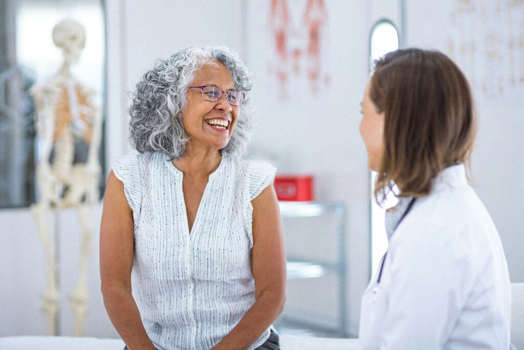 An older woman with gray curly hair smiles while talking to a female doctor in a white coat during a medical appointment in a bright Twin Cities exam room. A skeleton model is visible in the background.