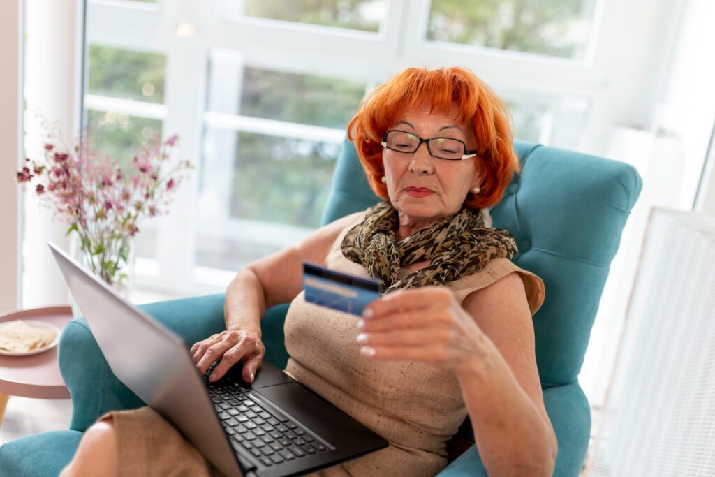 An older woman with red hair and glasses sits in a blue armchair using a laptop. Holding a credit card, she appears focused—perhaps researching estate planning in Minnesota. Bright windows illuminate the room in the background.