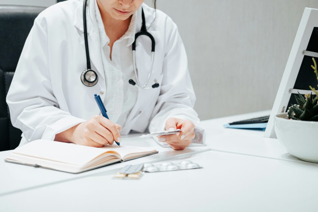 A doctor in a white coat with a stethoscope writes in a notebook while holding a pack of pills, ready to discuss the health care proxy role at a desk with medication and a plant nearby.