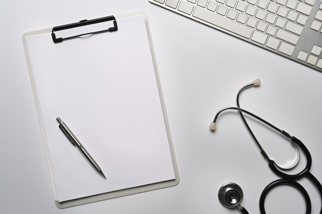 A blank clipboard with a pen, a stethoscope, and part of a computer keyboard are arranged on a white desk, ready for drafting a Health Care Directive.