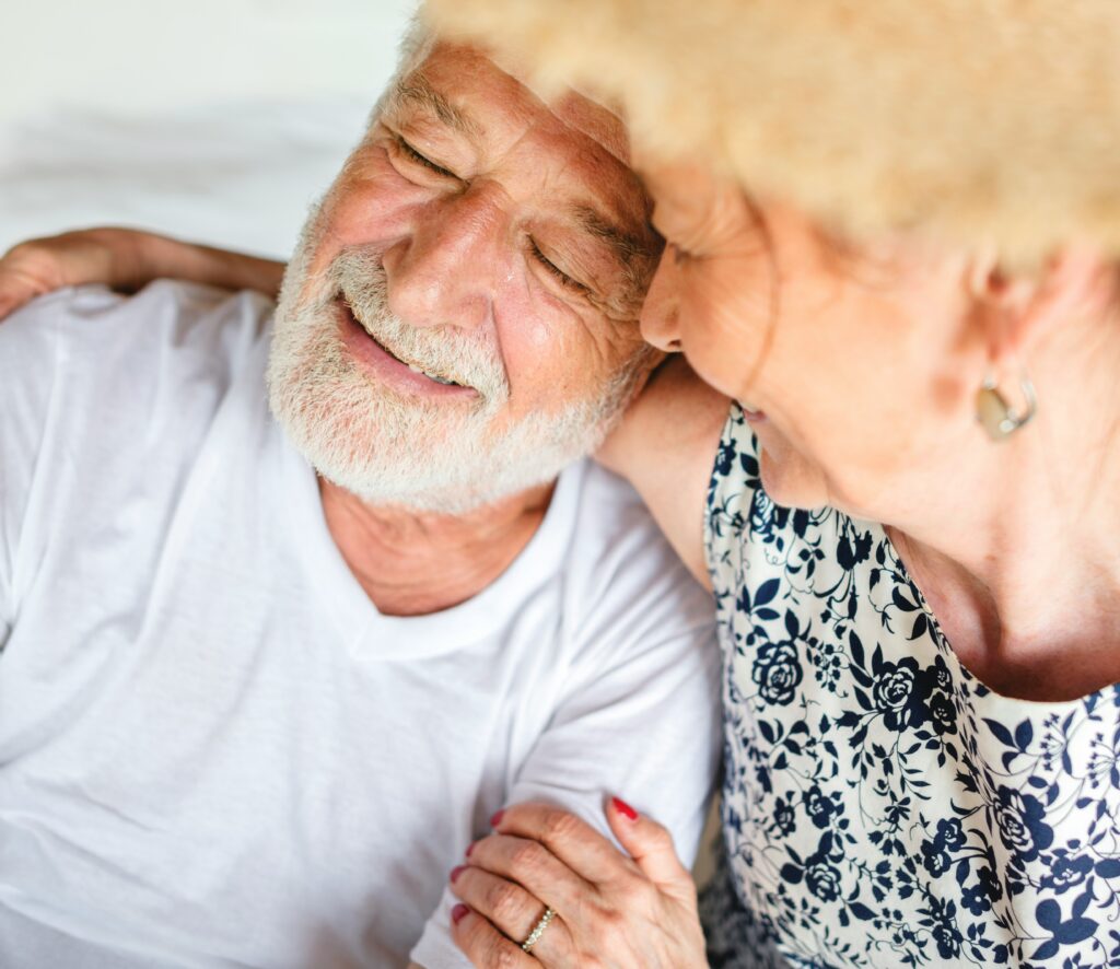 An elderly couple sits close together, smiling warmly in their Twin Cities home. The man wears a white t-shirt and the woman is in a floral dress with her hand on his arm, reflecting affection, happiness, and thoughtful life care planning.