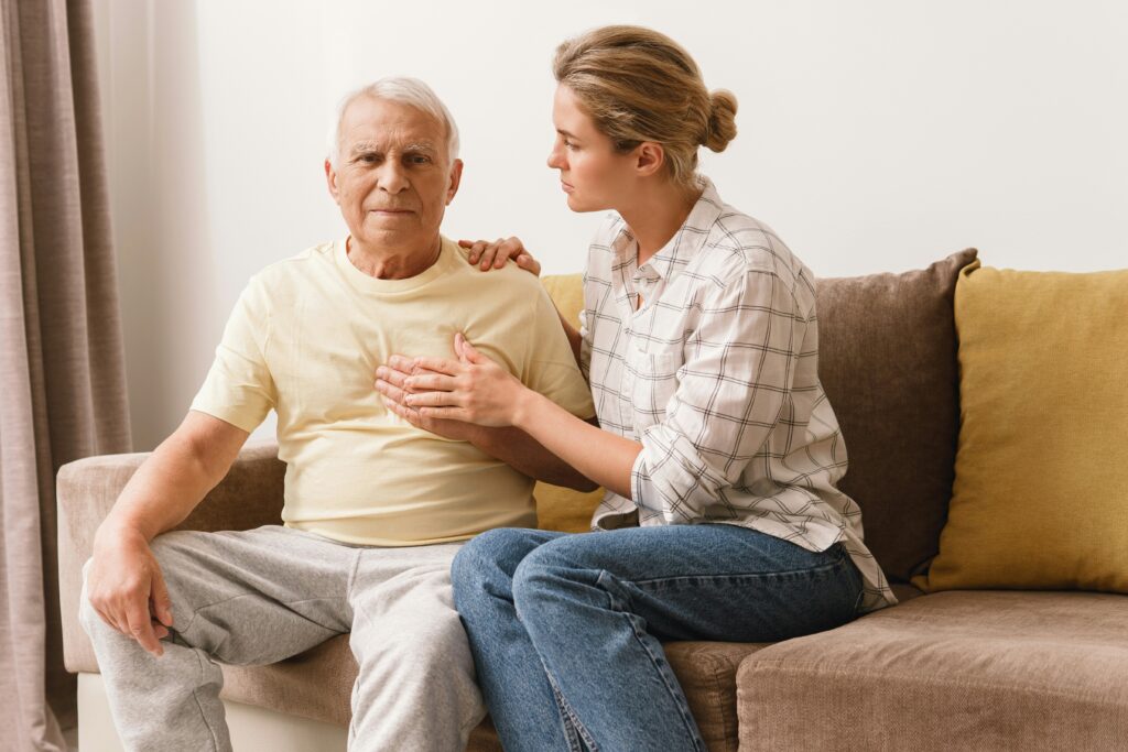 A young woman sits beside an older man on a couch, gently touching his chest and shoulder, showing concern and support—perhaps discussing important matters like Medicaid planning or Minnesota elder law. The man looks serious while the woman appears caring and attentive.