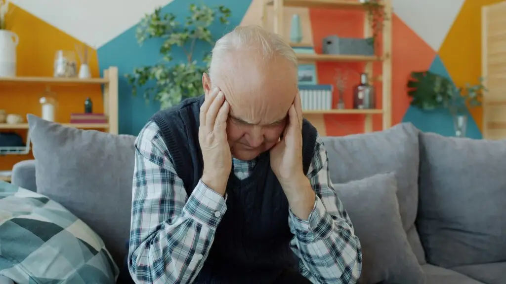 Older man on a couch holding his head in his hands