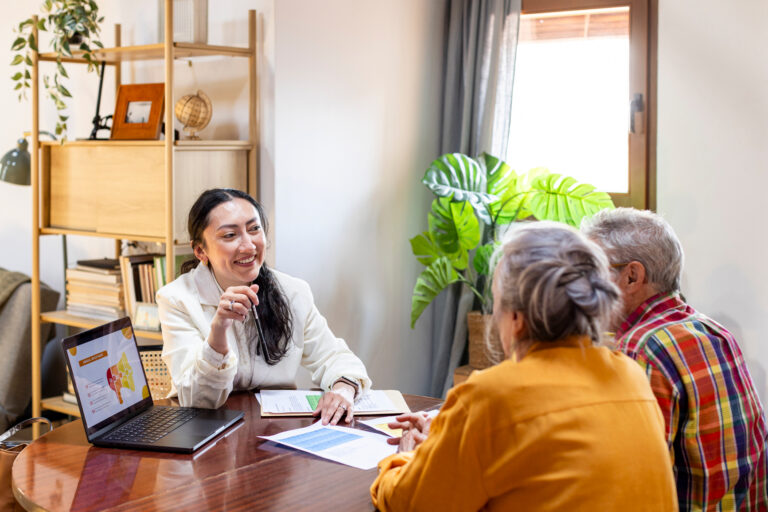 A woman sits at a round table talking to two older adults about estate planning Minnesota. She smiles and gestures with a pen while a laptop and papers are on the table in a cozy, well-lit room with a large houseplant by the window.