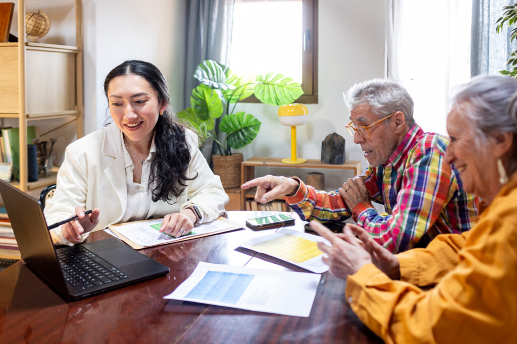 A young woman explains Medicaid planning on a laptop to two older adults sitting at a table with papers and a smartphone. All three are smiling and engaged in conversation in a bright, cozy Twin Cities room.