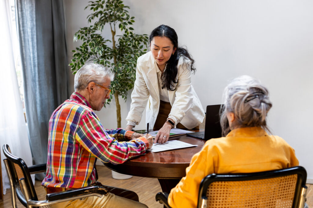 A woman in a white blazer stands beside a table, assisting two older adults with estate planning Minnesota documents and paperwork in a bright room with a plant in the background.