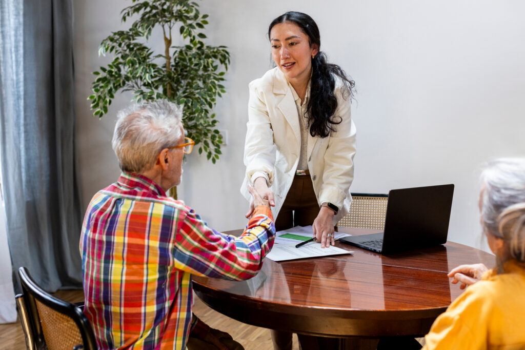 A woman in a white blazer, possibly an elder law attorney, smiles and shakes hands with an older man in a plaid shirt at a round table in the Twin Cities, with a laptop and Medicaid planning documents nearby. Another older person is seated close.