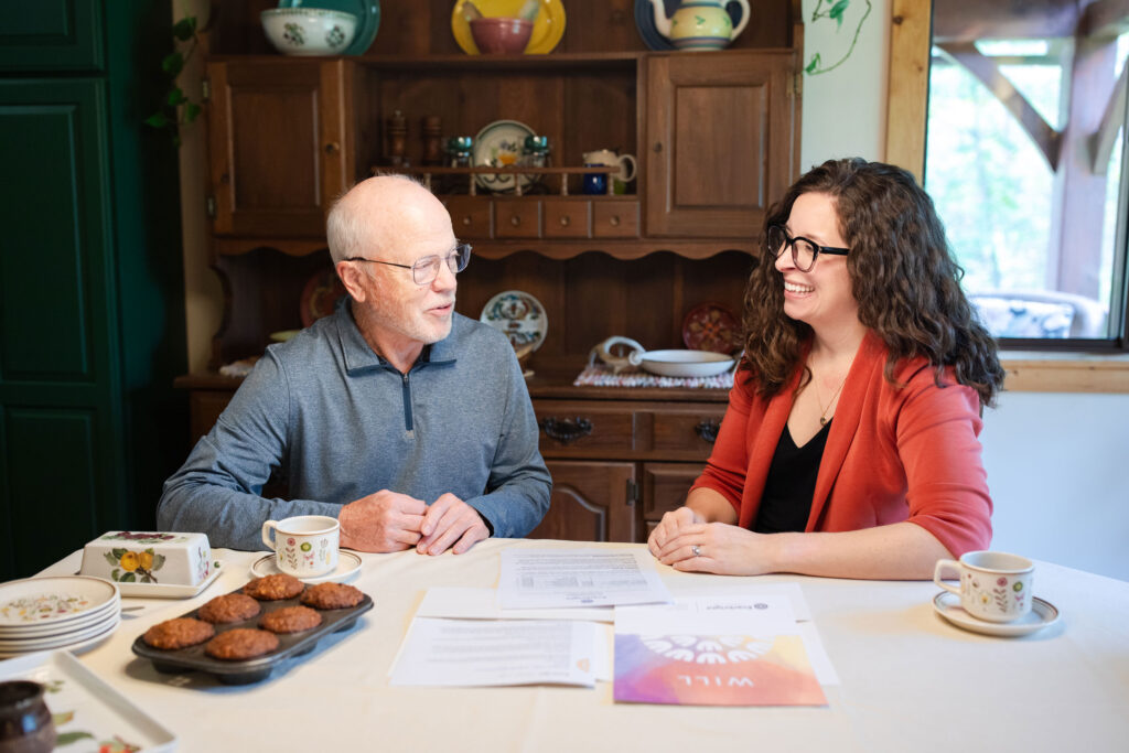 An older man and a younger woman sit at a table covered with papers—some labeled WILL—coffee cups, and muffins, smiling as they discuss life care planning in a cozy, warmly lit Twin Cities kitchen.