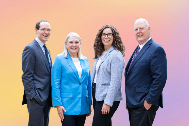 Four professionally dressed adults—three women and one man—stand in a row, smiling, against a pastel gradient background of pink, yellow, and orange hues, representing trusted elder law attorney services in the Twin Cities.