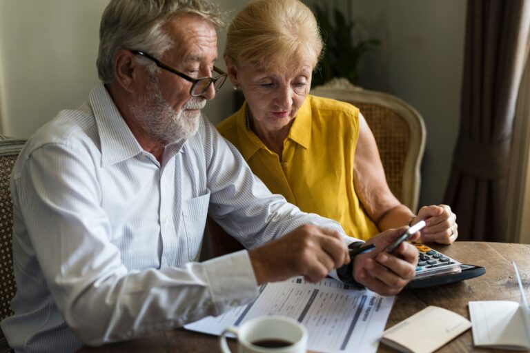 Elderly man and woman sitting at table reviewing documents. Man pointing at smartphone while woman holds calculator. Papers and coffee on table.