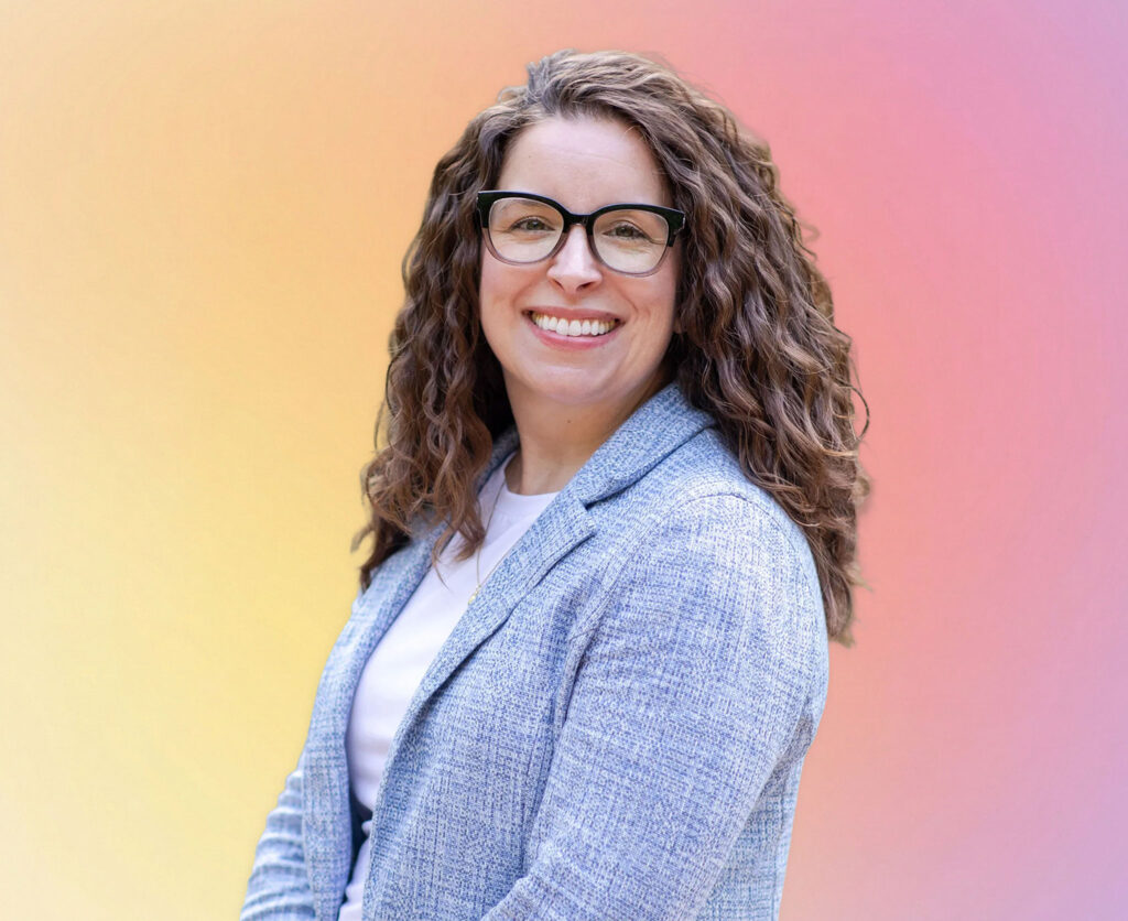 A woman with long curly brown hair and glasses smiles while wearing a light gray blazer over a white top, standing in front of a gradient yellow-to-pink background—reflecting the approachable spirit of an estate planning Minnesota attorney.