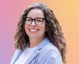 Lauren Fink wearing a light gray blazer over a white shirt, standing in front of a pink and yellow gradient background.