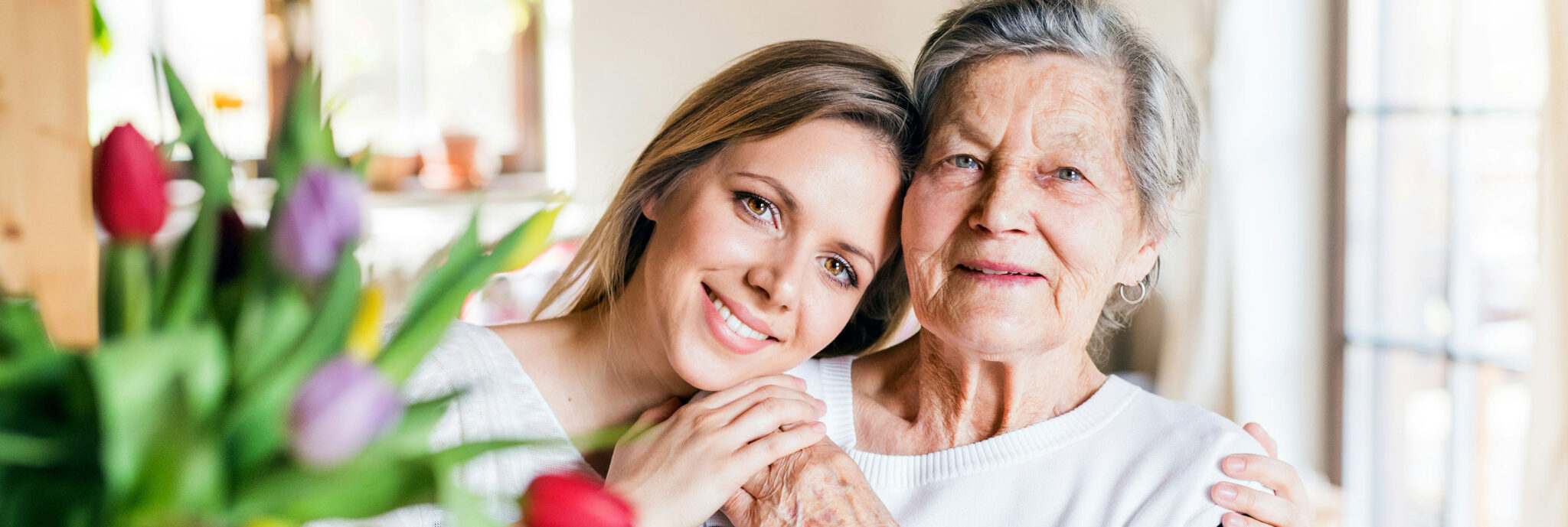 A young woman smiles and embraces an elderly woman, both in white tops, with a vase of colorful tulips in the foreground—capturing the warmth and connection that estate planning Minnesota specialists help families preserve.