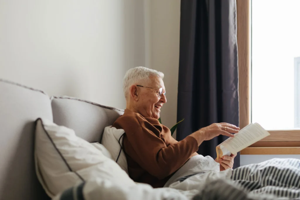 Older man smiling while reading a book in bed
