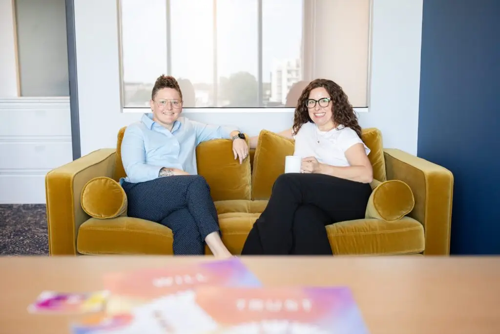 Cassie and Lauren sit and smile on a mustard yellow sofa in a bright, modern office. Both wear glasses and business-casual attire. Colorful blurred papers rest on a wooden table in the foreground.
