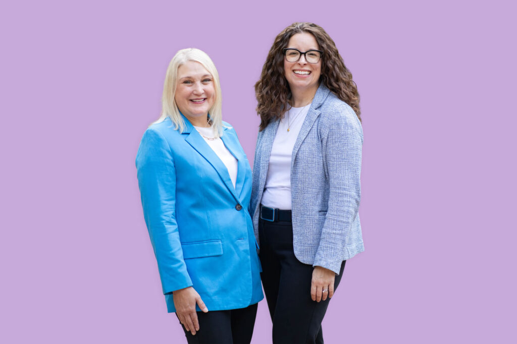 Two women standing side by side, both smiling and wearing blazers, against a plain light purple background—perfect for promoting life care planning or Medicaid planning services in the Twin Cities.