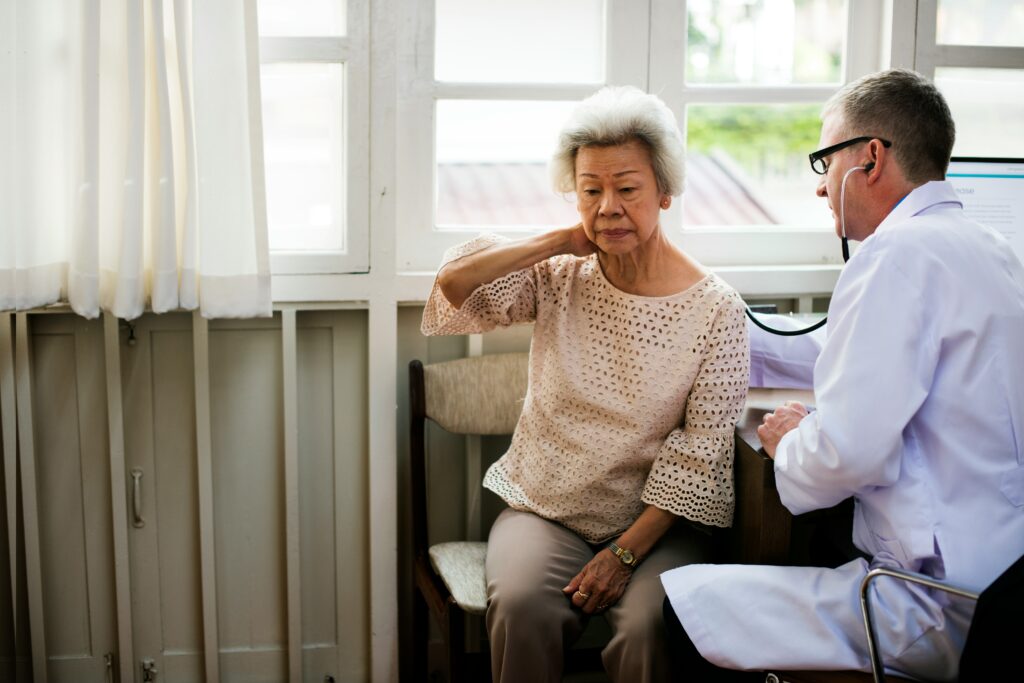 An elderly woman sits on a chair, touching her neck and looking uncomfortable, while a doctor—possibly discussing Medicare options—listens to her back with a stethoscope in a bright room with large windows.