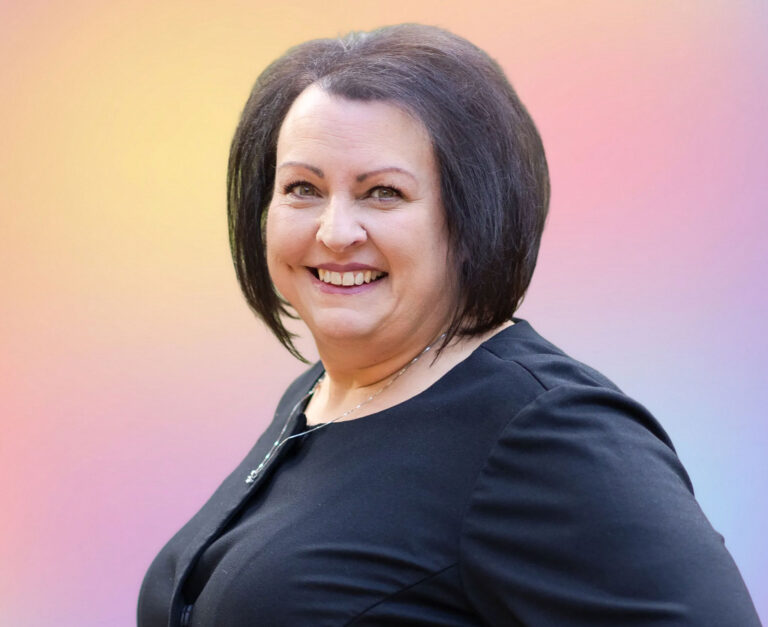 A woman with short dark hair and a black top smiles at the camera, standing in front of a soft, pastel-colored gradient background—perfect for professional profiles in life care planning or estate planning Minnesota services.