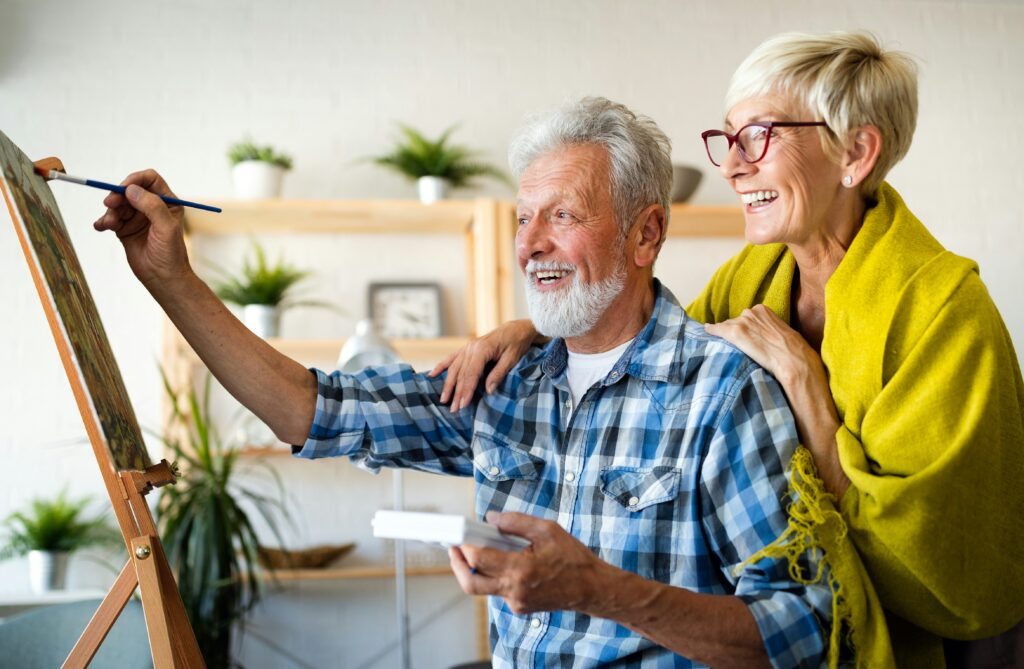 An older man with a white beard paints on a canvas, smiling, while a woman with short blonde hair and glasses stands beside him, laughing with her hands on his shoulders. They appear happy and relaxed in their bright Twin Cities home.