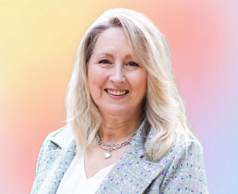 A smiling woman with long blonde hair wearing a light patterned blazer and layered necklaces, standing in front of a soft pastel gradient background—an approachable expert in estate planning Minnesota.