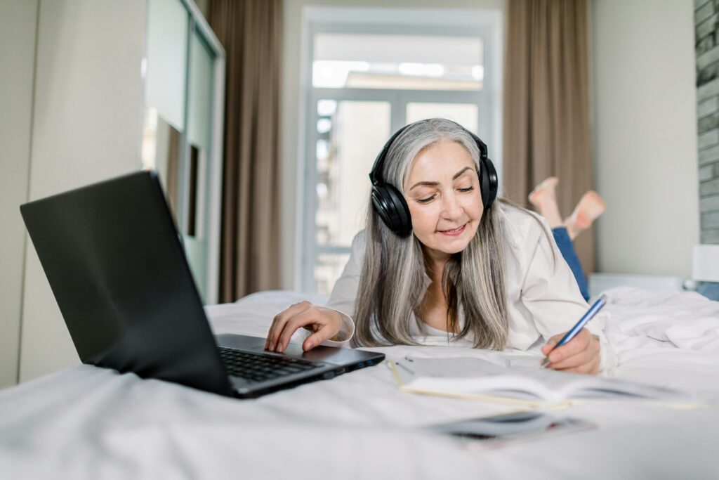 A woman with gray hair wearing headphones lies on a bed, smiling while writing in a notebook and using a laptop, planning her future with estate planning Minnesota resources, with a bright window in the background.