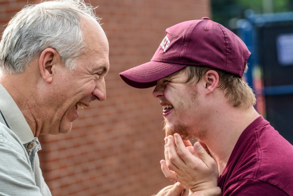 An older man and a young man wearing a maroon cap smile and laugh together outdoors, sharing a joyful, affectionate moment. A red brick wall is in the background—the perfect scene for discussing life care planning in the Twin Cities.