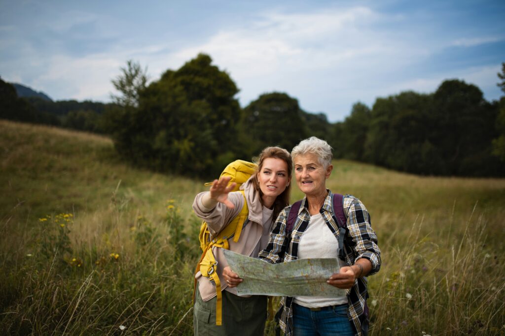 Two women with backpacks stand in a grassy field; one points ahead while the other holds a map, appearing to be navigating—much like life care planning for the future. Trees and a cloudy sky form the backdrop.