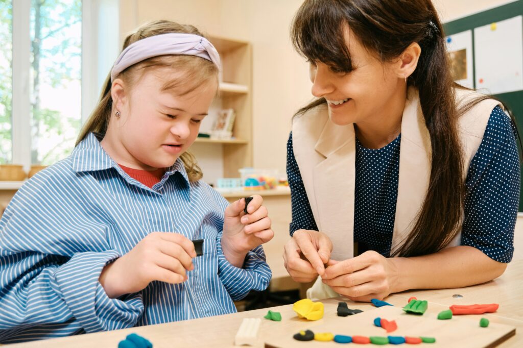 A young girl with Down syndrome and an adult woman smile and shape colorful modeling clay together at a table in a bright, cheerful room, highlighting the caring support often fostered by Minnesota elder law professionals.