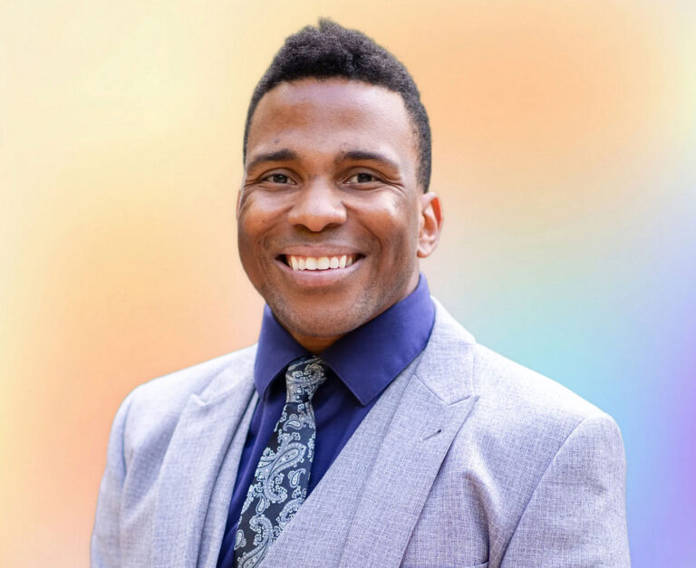 A man with short hair, wearing a light gray suit, a dark blue shirt, and a patterned tie, smiles in front of a softly blended pastel background—exuding the professionalism of an elder law attorney specializing in estate planning Minnesota.