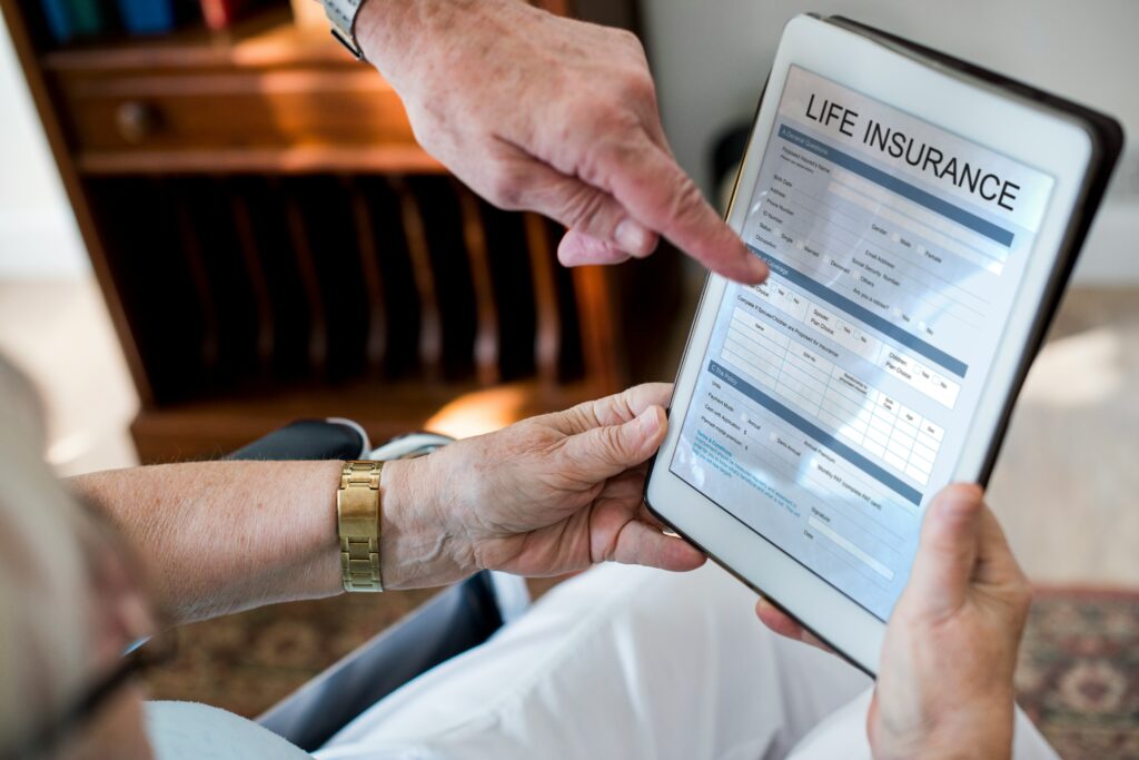 An elderly person holds a tablet displaying a life insurance form while another person, possibly an elder law attorney, points at the screen to assist with the application process. A wooden cabinet is visible in the background.