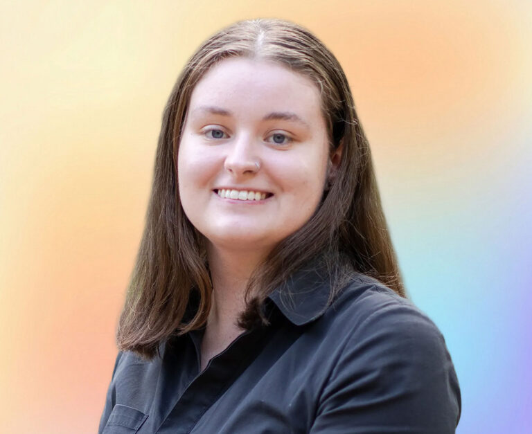 A young woman with straight brown hair and fair skin smiles at the camera. She is wearing a dark collared shirt, standing against a soft, colorful gradient background—an approachable Minnesota elder law professional ready to help with your life care planning.