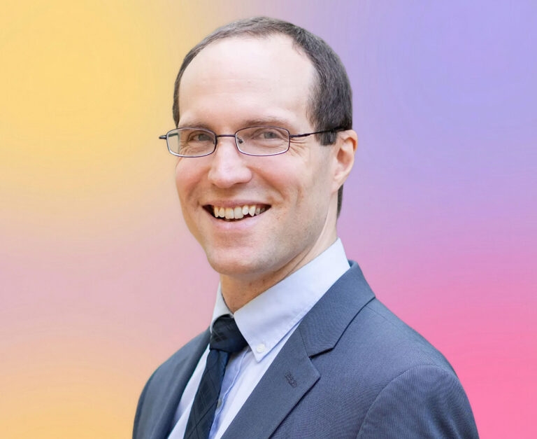 A man wearing glasses, a suit, and a tie is smiling at the camera. The background is a gradient of yellow, purple, and pink colors, suggesting he may be a Minnesota elder law attorney ready to assist with legal needs.