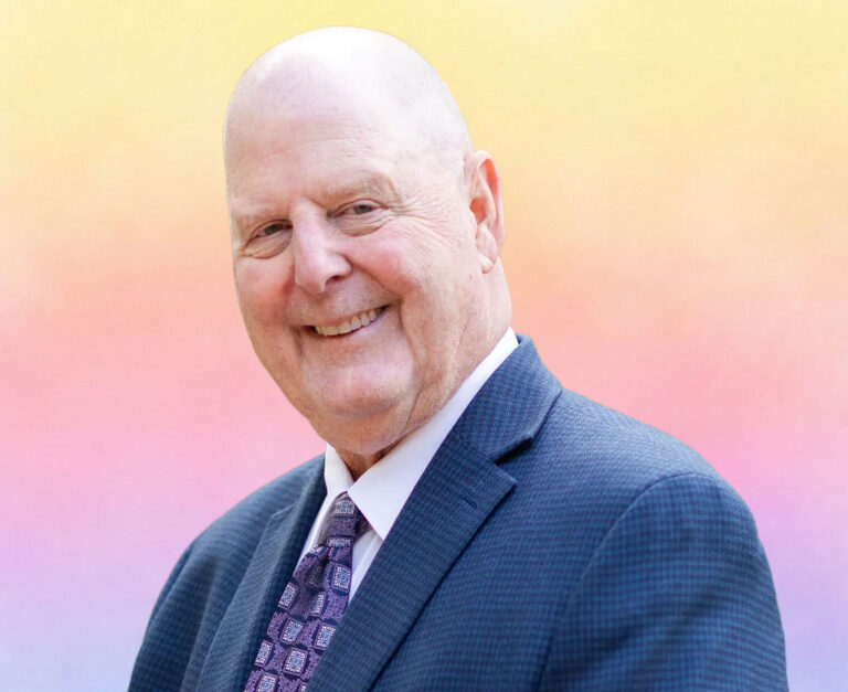 A smiling bald man in a dark suit and patterned tie stands before a soft gradient background of yellow, pink, and purple tones—reflecting his expertise in Minnesota elder law and Medicaid planning for the Twin Cities community.