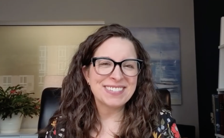 A woman with long curly brown hair and black glasses smiles at the camera. She is sitting indoors in front of a blurred painting and a plant, wearing a dark floral top—reflecting her thoughtful approach to Life Care Planning.