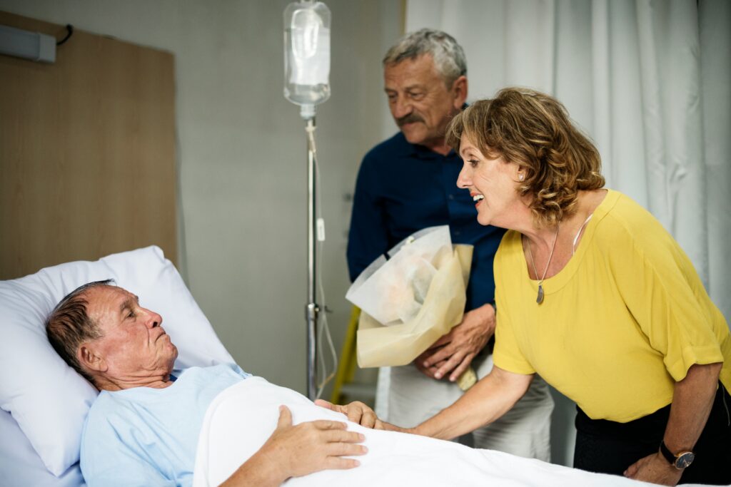 An elderly man lies in a hospital bed while a woman in a yellow shirt smiles and holds his hand. Another man, possibly involved in estate planning or trust law decisions, stands beside her holding a bouquet. An IV drip is visible next to the bed.