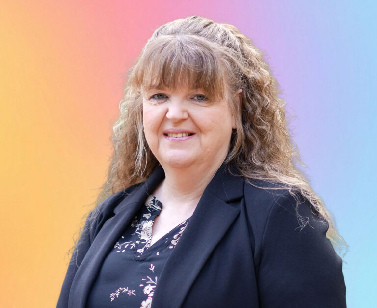 A woman with long, wavy light brown hair and bangs, wearing a black blazer over a floral blouse, smiles in front of a colorful gradient background—reflecting her dedication to Minnesota elder law and Medicaid planning in the Twin Cities.
