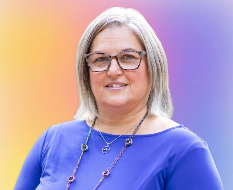 A woman with shoulder-length gray hair and glasses smiles at the camera. She is wearing a blue top and a necklace. The vibrant gradient background reflects her expertise in life care planning for the Twin Cities community.
