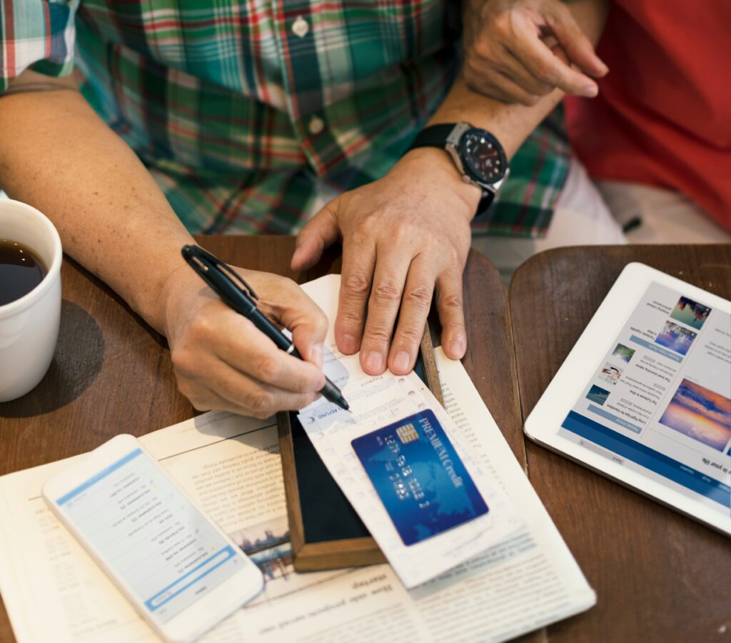 A person writes a check on a table with a credit card, open checkbook, smartphone, tablet, cup of coffee, and another person’s hand resting on their arm—capturing a thoughtful moment in Minnesota elder law or life care planning.
