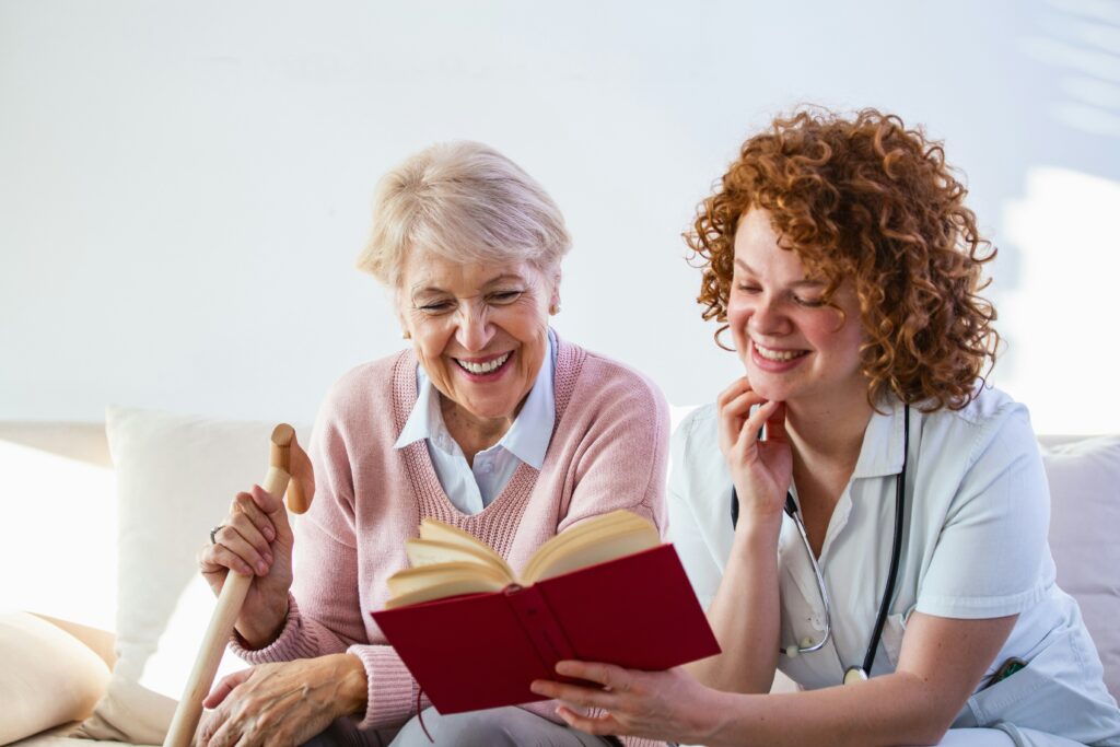 An older woman with a cane and a younger woman in a medical uniform sit together on a couch, smiling and reading a red book—illustrating the caring support offered by Minnesota elder law professionals.