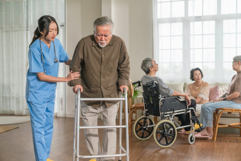 A nurse assists an elderly man using a walker, while three other seniors sit and converse in the background of a bright, communal living space—reflecting the caring environment supported by Minnesota elder law and life care planning.
