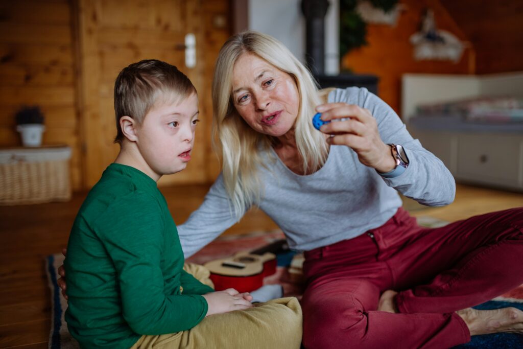A woman and a young boy with Down syndrome sit on the floor indoors, focused and engaged as she shows him a small blue object. This moment highlights the importance of understanding resources like the ABLE Age Adjustment Act Fact Sheet.
