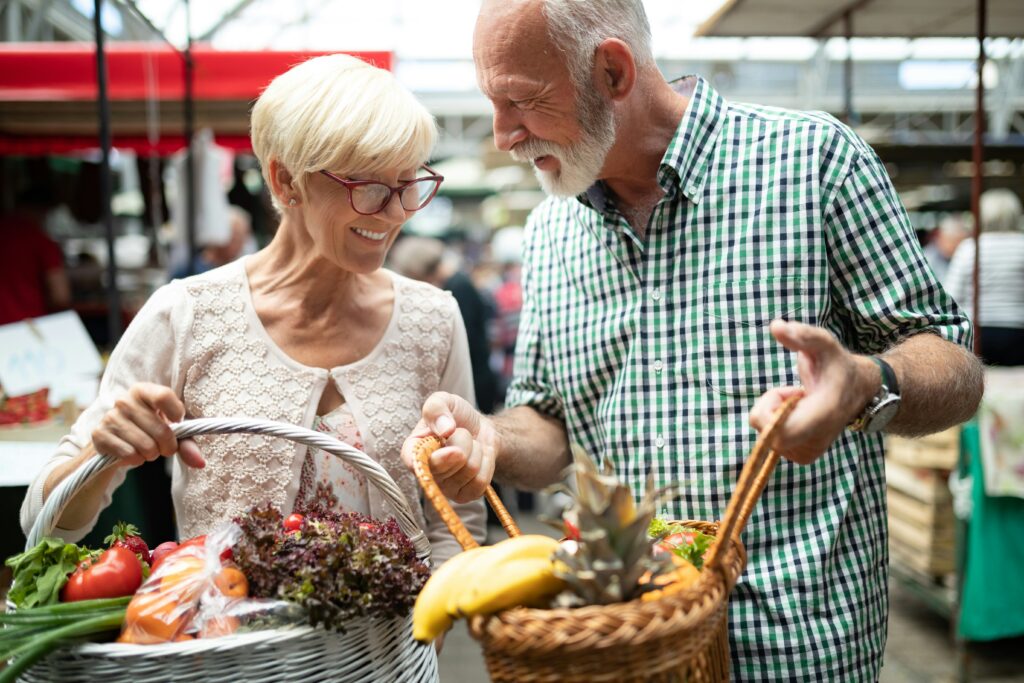 An older couple smiles at each other while holding baskets filled with fresh fruits and vegetables at an outdoor market—a joyful moment that estate planning Minnesota can help protect for generations to come.