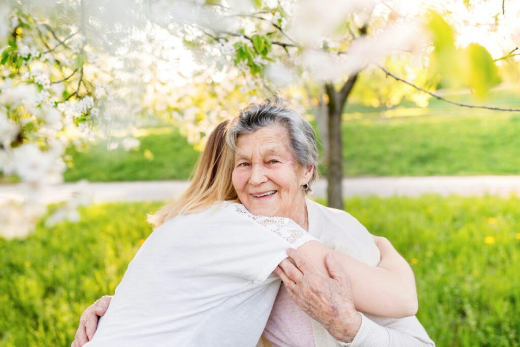 An elderly woman smiles warmly while hugging a younger person outdoors in Minnesota, surrounded by blooming trees and green grass—a perfect moment of family connection and Supported Decision-Making.