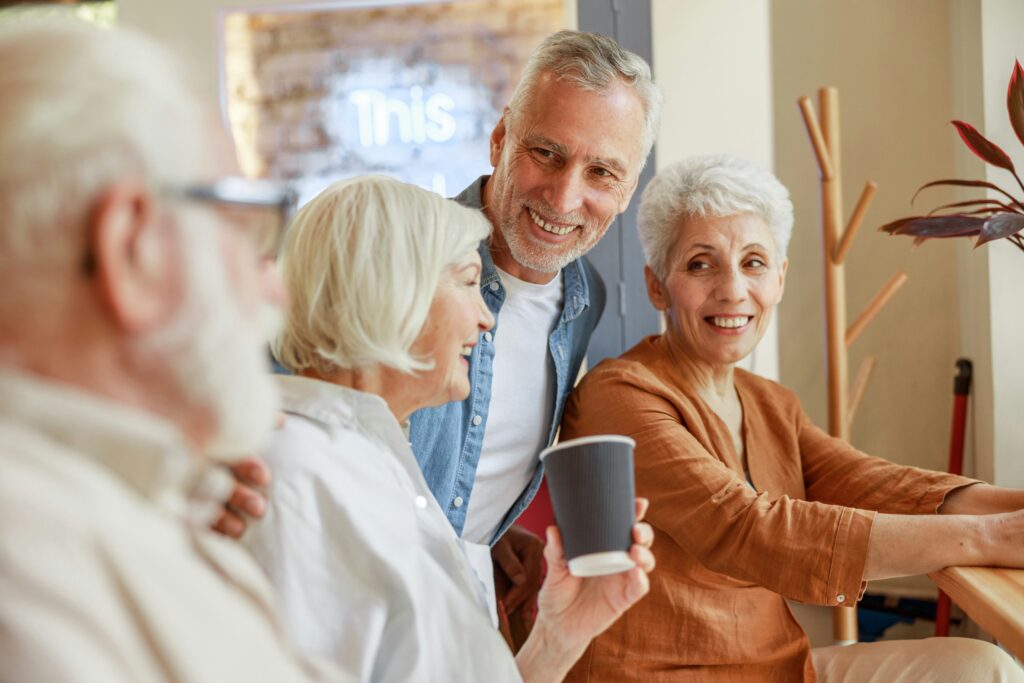 Four older adults are sitting together indoors, smiling and talking. One woman holds a paper cup, and a man stands behind her, smiling warmly at the group as they discuss legal tools like guardianship for Minnesota families. The atmosphere is cheerful and friendly.