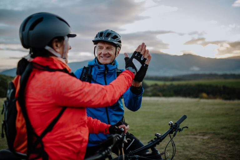 Two cyclists wearing helmets and jackets give each other a high five while smiling outdoors, celebrating getting rights back, with bikes in front of them and mountains visible in the background at sunset.